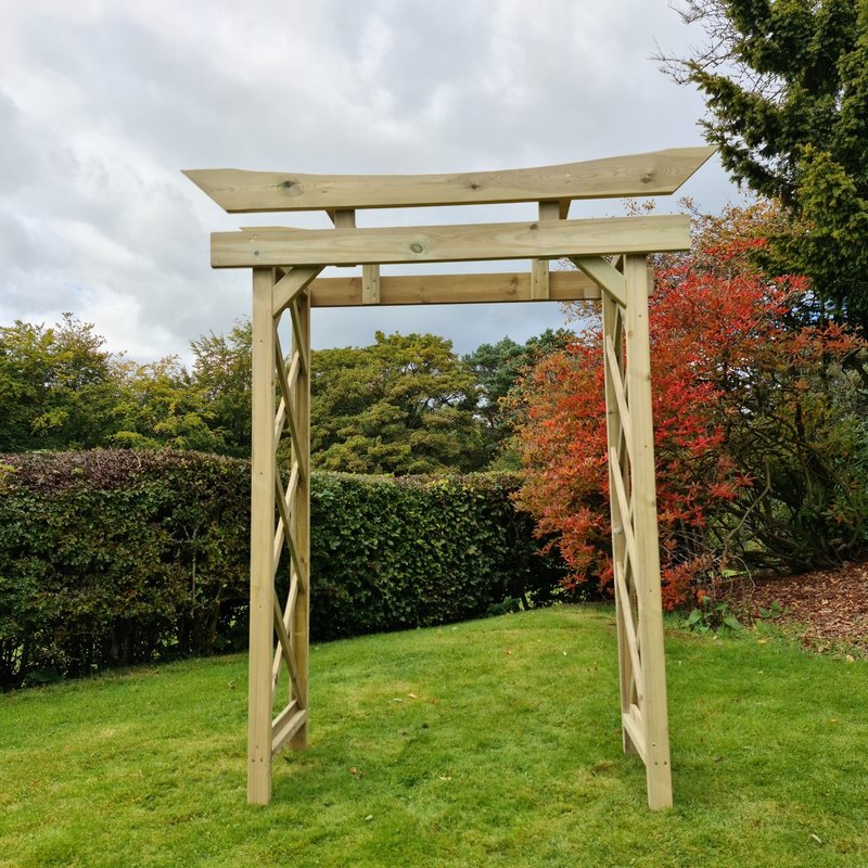 A light natural wood garden archway with twisted side supports, set on a green lawn with hedges, trees, and an overcast sky in the background.
