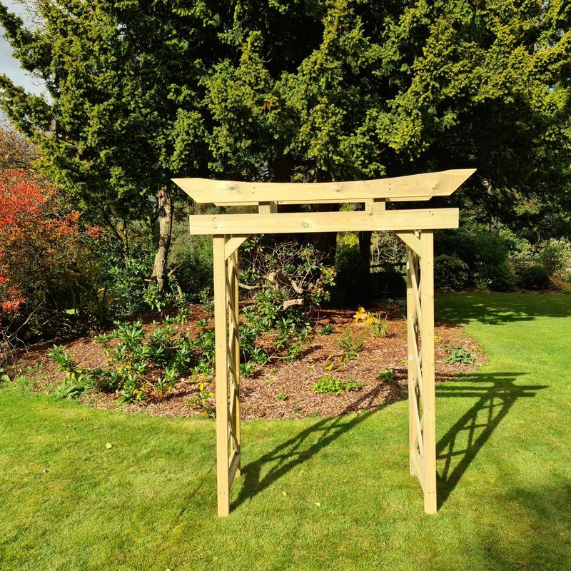A light natural wood garden archway with a curved top, standing on a green lawn in a landscaped garden with trees and mulch in the background.