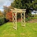 A light natural wood garden archway with a slatted roof, set on a green lawn in a backyard with trees and shrubs in the background.